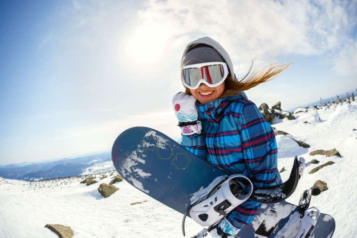 Frau in Wintersportklamotten und Snowboard in der Hand lächelt fröhlich 